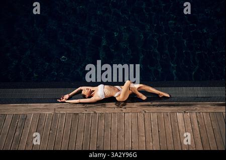 Sich hinlegen und das Sonnenlicht genießen. Frau in weißem Bikini ist in der Nähe des Schwimmbades. Stockfoto