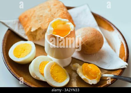 Gekochtes Ei in Eierbecher, in Schale und auf Teller geschnitten, Löffel und Brot (Brötchen), Handtuch., Nahaufnahme. Gesundes Frühstück. Stockfoto