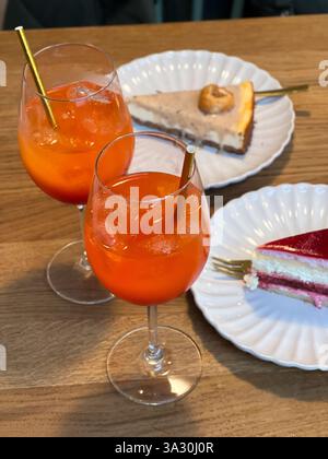 Nahaufnahme von zwei Orangen-Cocktail-Drinks und zwei Stück Kuchen auf weißen Tellern, Holztisch Stockfoto