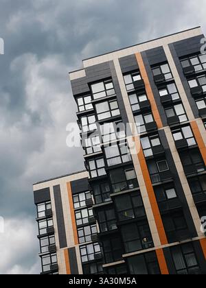 Modernes Wohngebäude vor bewölktem Himmel. Dunkelgraues Apartmentgebäude mit großen Fenstern und beigen n orangen Streifen. Stockfoto