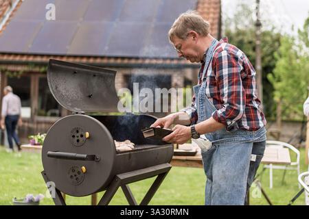 Senior Mann kocht auf einem Gartengrill, genießt Outdoor-Kochen und Freizeitaktivitäten. Reifer Mann im Overall, der im Garten Essen grillt, aktiv Stockfoto