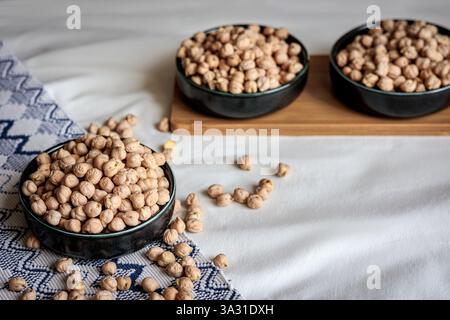 Getrocknete Kichererbsen in einer schwarzen Keramikschale auf einem gemusterten Textil mit verstreuten Hülsenfrüchten, die eine gemütliche und natürliche Präsentation von gesunder pflanzlicher Basis schaffen Stockfoto