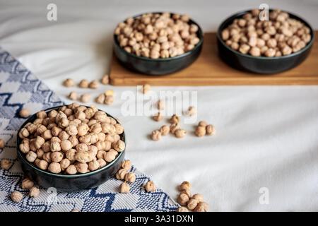 Getrocknete Kichererbsen in einer Keramikschale auf einem strukturierten Stoff mit verstreuten Kichererbsen und Holztablett im Hintergrund, das natürliche Lebensmittel und Rusti hervorhebt Stockfoto