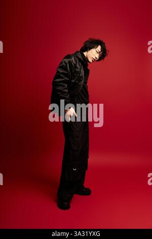 Ein junger asiatischer Mann in Schwarz posiert stilvoll in einem leuchtend roten Studio. Stockfoto