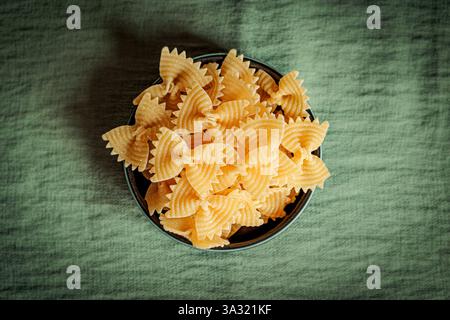 Blick von oben auf eine schwarze Keramikschale gefüllt mit ungekochten Farfalle-Pasta auf einem grünen Stoffhintergrund. Einfache und strukturierte Komposition, perfekt zum Kochen Stockfoto