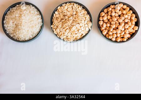 Drei Keramikschüsseln mit Hafer, Reis und Kichererbsen auf weißer Oberfläche, mit verstreuten Körnern und Hülsenfrüchten, die natürliche, rustikale Lebensmittelkompositionen schaffen Stockfoto