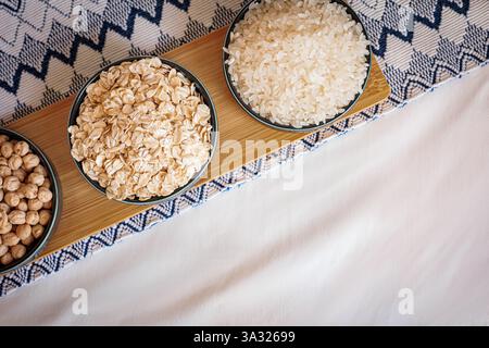 Von oben aus sehen Sie Schüsseln mit Hafer, Reis und Kichererbsen auf einem Holzbrett, die mit einem dekorativen blau-weißen Tuch auf einer weißen Oberfläche platziert werden. A Stockfoto