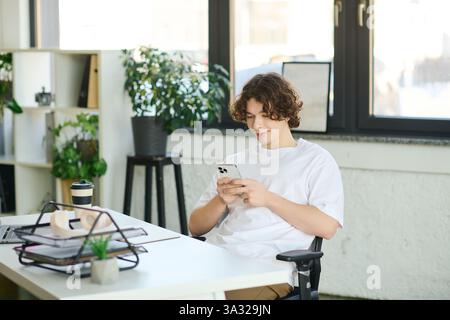 Ein hübscher junger Mann sitzt bequem in einem Bürostuhl, konzentriert sich auf sein Smartphone und lächelt. Stockfoto