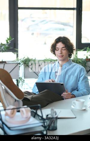 Ein gutaussehender junger Profi sitzt bequem an seinem Schreibtisch und konzentriert sich auf sein Tablet. Stockfoto