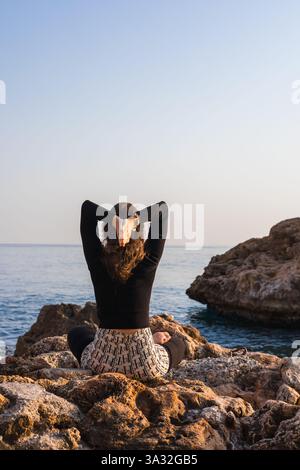 Eine Frau sitzt im Kreuz auf einem großen Felsen am Meer oder Meer und übt Yoga in einem Sonnenuntergang im Freien. Die ruhige Szene zeigt Berge in d Stockfoto