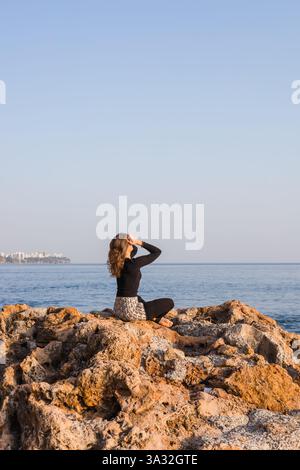 Eine Frau sitzt auf einer Felsformation am Meer oder Ozean und blickt in die Ferne. Die helle Sonne scheint auf das friedliche Wasser und schafft eine ruhige Atmosphäre Stockfoto