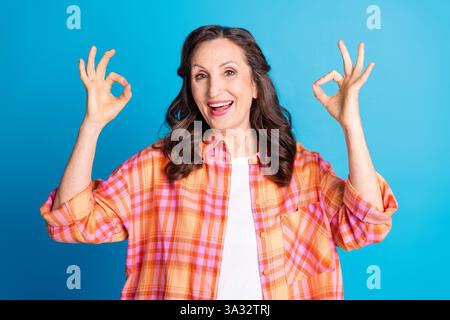 Lächelnde ältere Frau mit Gesten von OK-Zeichen in stilvollem kariertem Hemd vor blauem Hintergrund Stockfoto