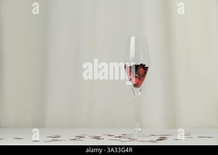 Weinglas mit Herzkonfetti auf cremefarbenem Hintergrund. Minimalistisches Urlaubsfoto zum Valentinstag Stockfoto
