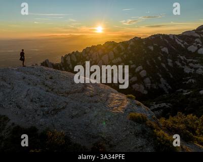 Eine Einzelfigur steht auf einer felsigen Klippe in Montserrat, Katalonien, während die Sonne aufgeht und warmes Licht über die zerklüftete Landschaft und die fernen Hügel wirft. Stockfoto