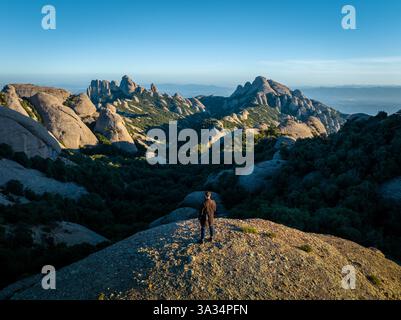 Eine Person steht auf einem felsigen Hügel mit Blick auf die atemberaubenden Montserrat Berge in Katalonien, Spanien, mit klarem blauen Himmel, der einen atemberaubenden Pano erzeugt Stockfoto