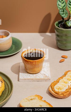 Eine gemütliche Frühstücksszene mit einer keramischen Tasse schwarzen Kaffee, Toast mit Erdnussbutter und Bananenscheiben, Mandeln und Topfpflanze in einem warmen, invit Stockfoto