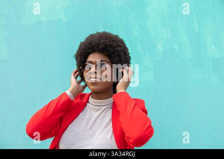 Stilvolle Frau in leuchtend roter Jacke steht vor einem türkisfarbenen Hintergrund, passt ihre Sonnenbrille an und lächelt und verkörpert ein selbstbewusstes und modernes li Stockfoto