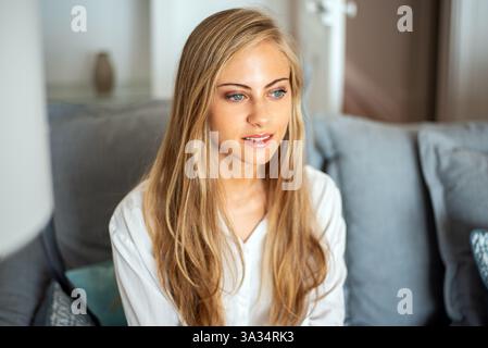 Eine ruhige Frau mit langen blonden Haaren sitzt auf einem bequemen grauen Sofa und blickt nachdenklich in die Ferne. Das gemütliche Zimmer bietet eine ruhige und einladende Atmosphäre Stockfoto