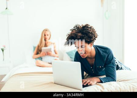 Ein junger Mann liegt mit einem Laptop auf einem Bett, während eine Frau mit einem Tablet im Hintergrund sitzt. Die Szene fängt ein gemütliches, technisches Ambiente in einem bri ein Stockfoto