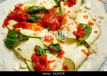Frische Zutaten wie geschnittene Zucchini, Basilikumblätter und Tomatensauce auf Pizzateig, die den Prozess der Zubereitung einer Pizza veranschaulichen. Stockfoto