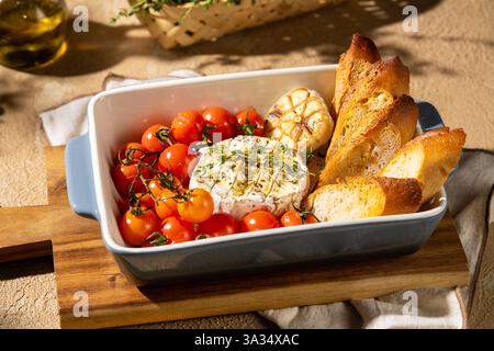 Ein herzhaftes Gericht mit gebackenem Camembert-Käse, garniert mit Kräutern, umgeben von gerösteten Kirschtomaten, Knoblauch und knusprigem Brot, serviert in einer cera Stockfoto