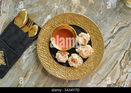 Von oben elegante Präsentation von Knödeln, serviert auf einem goldfarbenen Teller, gepaart mit einer scharfen roten Sauce, auf einer marmorierten Oberfläche. Die Einrichtung umfasst Stockfoto