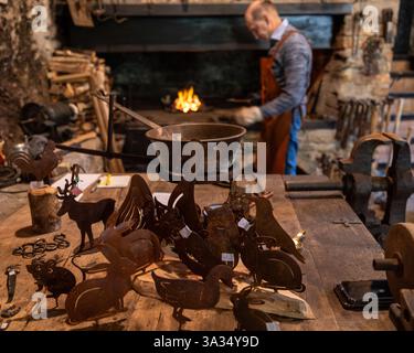 Ein Schmied arbeitet in der Nähe einer Schmiede in einer rustikalen Werkstatt mit handgefertigten Tierfiguren aus Metall auf einem Holztisch. Das warme Feuerlicht hebt das hervor Stockfoto