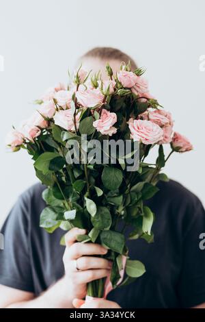 Ein junger Mann hält einen großen Strauß frischer rosafarbener Rosen und versteckt sein Gesicht dahinter. Der Hintergrund ist ein einfacher, neutraler Farbton, der das lebhafte betont Stockfoto