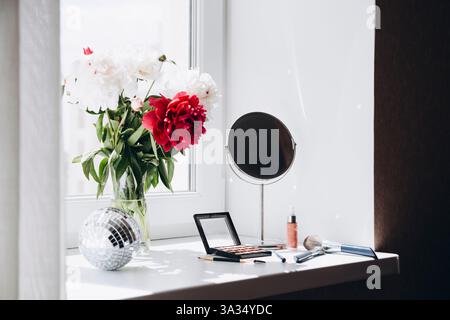 Ein lebhafter Blumenstrauß aus weißen und roten Pfingstrosenblüten in einer klaren Vase auf einer lichtdurchfluteten Fensterbank, begleitet von Make-up-Artikeln und einem runden Spiegel Stockfoto