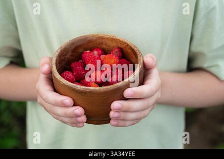 Nahaufnahme eines Kindes mit einer Holzschale gefüllt mit Reifen roten Erdbeeren, die Frische und Einfachheit vermittelt. Das blassgrüne Hemd des Kindes fügt eine kleine Note hinzu Stockfoto
