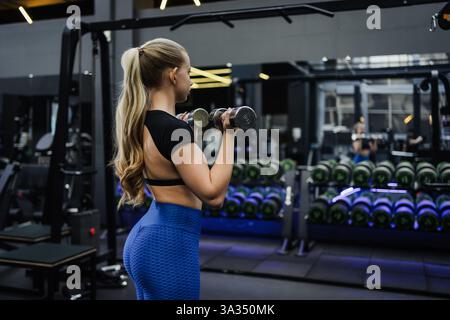 Eine blonde Frau mit langen Haaren, die mit Kurzhanteln in einem modernen Fitnessstudio arbeitet, umgeben von Fitnessgeräten, mit Stärke und Entschlossenheit, in s Stockfoto