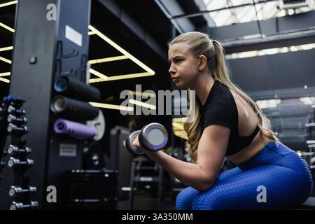 Eine konzentrierte blonde Frau mit blondem Haar führt Bizepslocken in einem modernen Fitnessstudio aus. Sie trägt ein schwarzes Oberteil und blaue Leggings, umgeben von Fitness Stockfoto