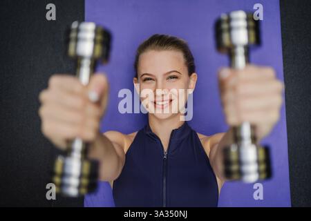 Blonde Frau, die mit Kurzhanteln trainiert, lächelnd und selbstbewusst auf einer lila Yogamatte. Eine perfekte Darstellung eines gesunden Lebensstils, Stärke und Abschreckung Stockfoto