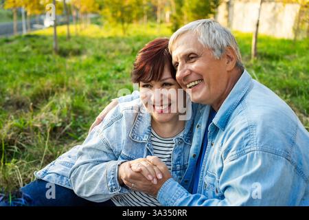 Ein fröhliches Seniorenpaar mit Jeansjacken, dicht im Park. Sie lächeln herzlich, umarmen sich gegenseitig, umgeben von üppigem grünem Gras und t Stockfoto