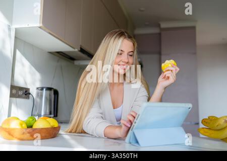Frau in einer gemütlichen Küche mit einer Tablette, die ein Stück Obst hält. Natürliches Licht hellt den Raum auf. Eine Schüssel mit Äpfeln und Orangen sitzt auf einem sauberen Topf Stockfoto