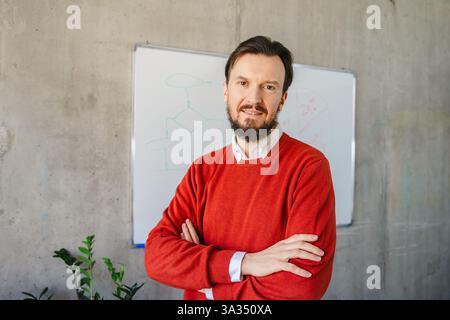 Ein Mann in einem roten Pullover steht mit überkreuzten Armen vor einem Whiteboard in einer modernen Büroumgebung und strahlt Professionalität und Führungsstärke aus. Stockfoto