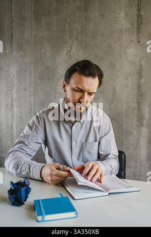 Ein Mann in einem Hemd sitzt an einem Tisch in einem minimalen Büro und schreibt in ein Notizbuch. Ein blaues Notizbuch und zerknittertes Papier sind in der Nähe, was seine nachdenkliche Untermalung unterstreicht Stockfoto