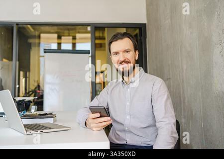 Ein Mann in einem hellen Hemd sitzt mit einem Smartphone an einem Schreibtisch. Ein Laptop und ein Notizblock begleiten ihn in einer hellen, modernen Büroumgebung. Stockfoto