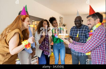 Eine fröhliche Gruppe von Leuten, die drinnen Geburtstag feiern. Sie tragen bunte Partyhüte und Leis und versammeln sich eifrig um einen Kuchen mit zündeter Kerze Stockfoto