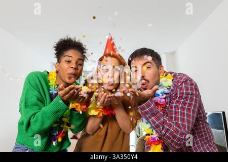 Drei Freunde in farbenfroher Kleidung und Leis blasen Konfetti und schaffen eine festliche Geburtstagsatmosphäre. Sie zeigen fröhliche Ausdrucksformen, die den Promi aufwerten Stockfoto