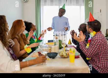 Eine vielfältige Gruppe von Familie und Freunden versammelt sich um einen Tisch und feiert einen Geburtstag mit Partyhüten, Getränken und Freude. Die Szene fängt Glück und ein Stockfoto