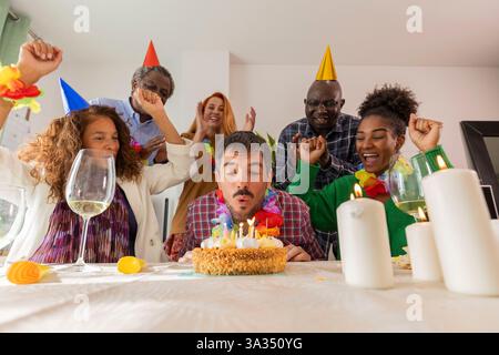Eine fröhliche Geburtstagsfeier mit Freunden, während die Geburtstagsperson Kerzen auf einem Kuchen ausbläst. Lachen, Partyhüte und festliche Dekorationen verstärken das Stockfoto