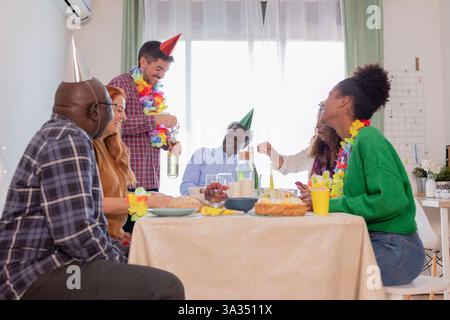 Eine Gruppe von Freunden, die zu Hause eine Geburtstagsfeier feiern. Sie tragen farbenfrohe Partyhüte und Leis, mit Getränken und Snacks auf dem Tisch, die Freude ausstrahlen Stockfoto