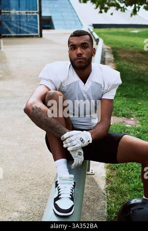 Ein junger American-Football-Spieler in einem weißen Trikot sitzt draußen auf einer Bank und strahlt Selbstvertrauen und Bereitschaft aus. Die Szene fängt das Wesen des Sports ein Stockfoto