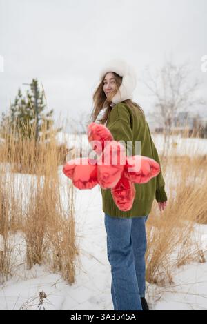 Eine Frau mit grünem Pullover und Pelzmütze hält in einem schneebedeckten Park rote Herzballons. Sie lächelt über ihre Schulter und nimmt den Winterbeobachter an Stockfoto
