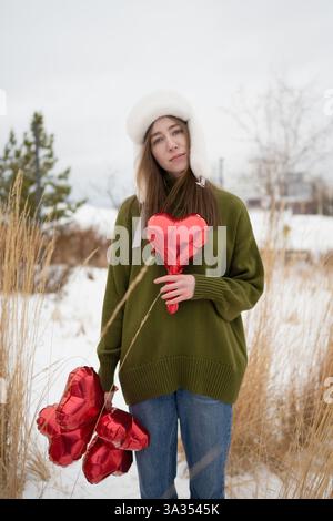 Frau in einem grünen Pullover, die rote Herzballons in einem verschneiten Park hält, umgeben von trockenem Gras, die Wärme und Liebe zur Kälte verkörpern. Stockfoto
