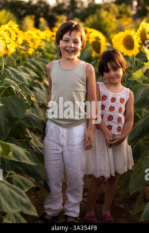 Zwei Kinder stehen fröhlich in einem Sonnenblumenfeld während der goldenen Stunde. Das warme Sonnenlicht hebt die leuchtend gelben Blüten und saftig grünen Blätter hervor, cr Stockfoto