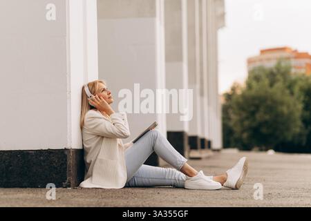 Eine Geschäftsfrau sitzt entspannt auf dem Boden und hört Musik über Kopfhörer. Sie lehnt sich an eine Gebäudemauer und genießt einen friedlichen Moment Stockfoto