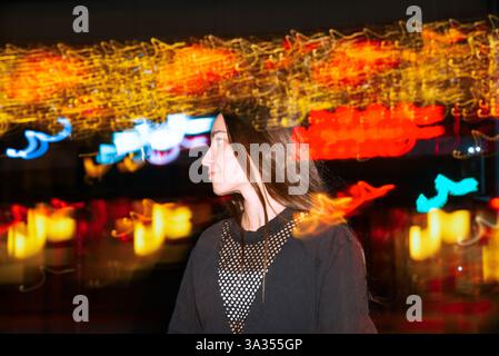 Eine Frau steht inmitten lebendiger Neonlichter mit Bewegungsunschärfe und schafft eine abstrakte und dynamische urbane Szene auf der Straße. Das Bild zeigt das Nachtleben e Stockfoto