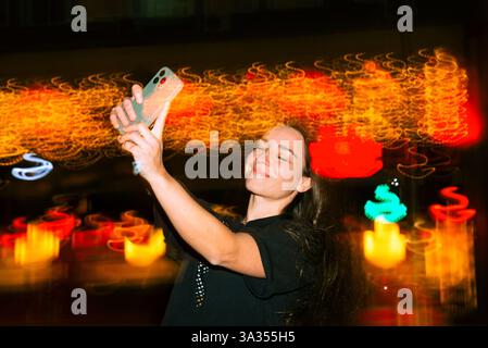 Eine Frau nimmt mit ihrem Handy ein Selfie in leuchtenden, bunten Lichtern der Stadt bei Nacht auf. Ihr Ausdruck ist fröhlich und spiegelt die lebhafte Atmosphäre in wider Stockfoto